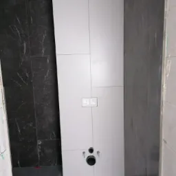 Montaż płytek w łazience w Koszalinie: białe i czarne płytki na ścianie, widoczne otwory na instalacje sanitarne i włączniki światła, czerwone krzyżyki dystansowe na podłodze.