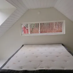 Poddasze z oknem, widok na las i czerwony domek, białe ściany i sufit z drewnianą boazerią, materac na podłodze z plamą.
