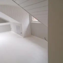 Białe, świeżo pomalowane poddasze z oknem w skosie dachu, widoczne pionowe panele ścienne i gładka podłoga, minimalistyczny design.