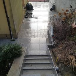 Kamieniarstwo Szymański Wojciech - Ułożony taras z szarych płytek, schody z szarego kamienia prowadzące w dół, widoczne narzędzia i materiały budowlane, krzewy i fragment ogrodu w tle.