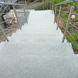 Kamieniarstwo Szymański Wojciech - Zewnętrzne schody z jasnoszarego kamienia z metalową balustradą, prowadzące w dół do ogrodu z tujami. Widok z góry, symetryczny kadr.