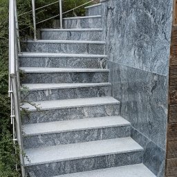 Kamieniarstwo Szymański Wojciech - Zewnętrzne schody z szarego kamienia z metalową balustradą, otoczone zielenią. Widoczne detale faktury kamienia i kostki brukowej u podstawy schodów.