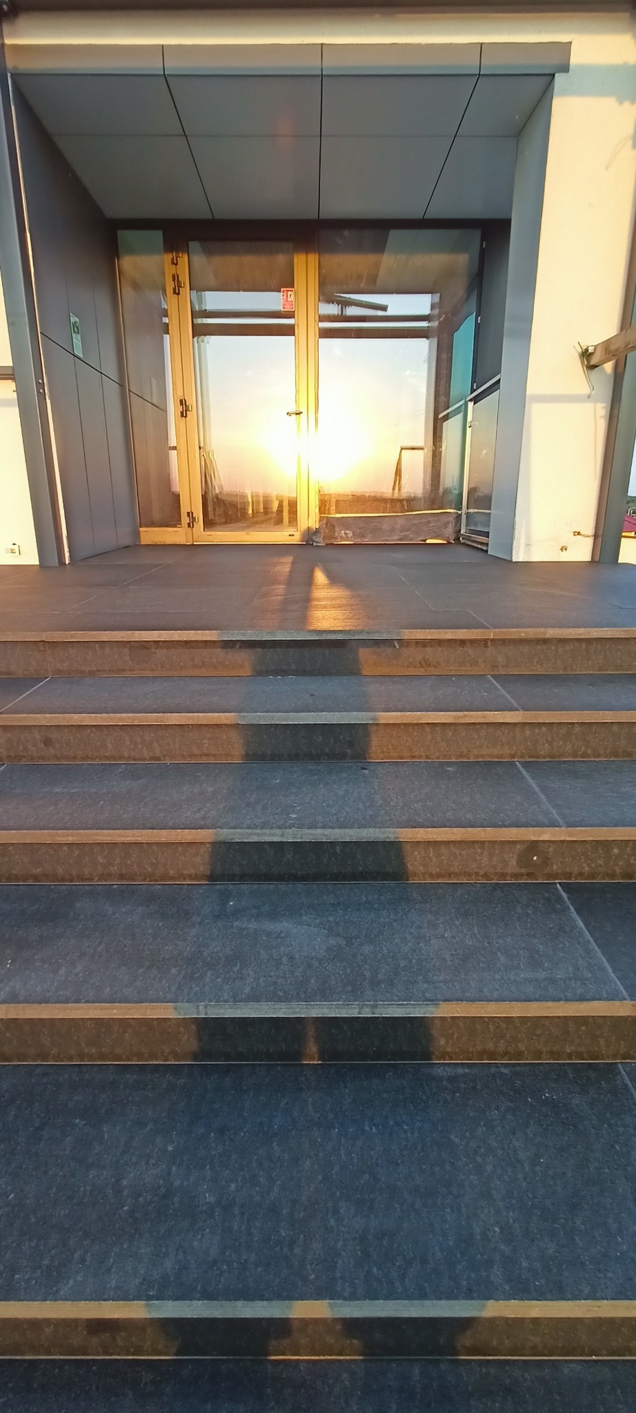Kamienne schody prowadzą do przeszklonego wejścia, w którym odbija się zachodzące słońce. Ciemne stopnie kontrastują z jasnym światłem w tle.