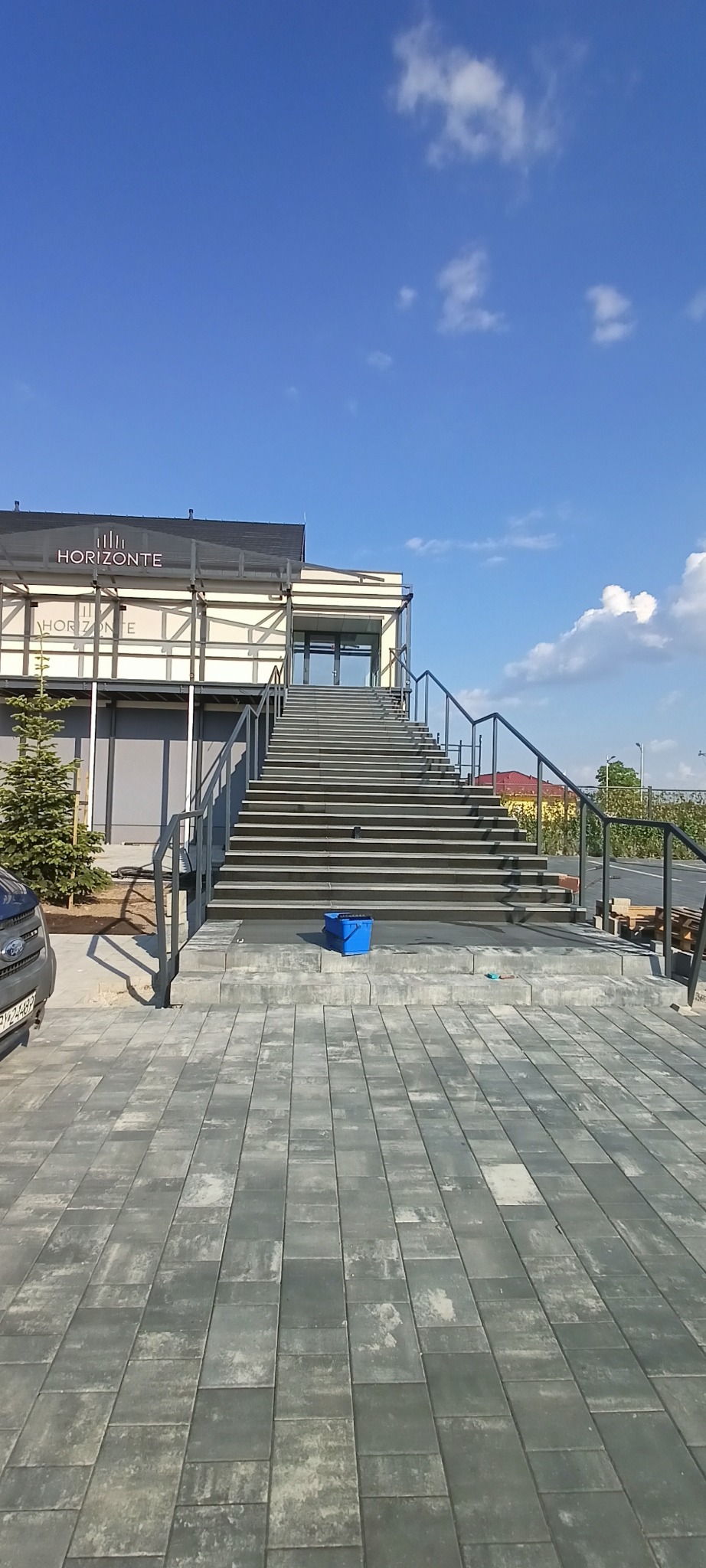 Zewnętrzne schody kamienne z szarych płyt prowadzące do budynku z napisem 'Horizonte', widoczne błękitne wiadro i fragment samochodu. Słoneczny dzień.