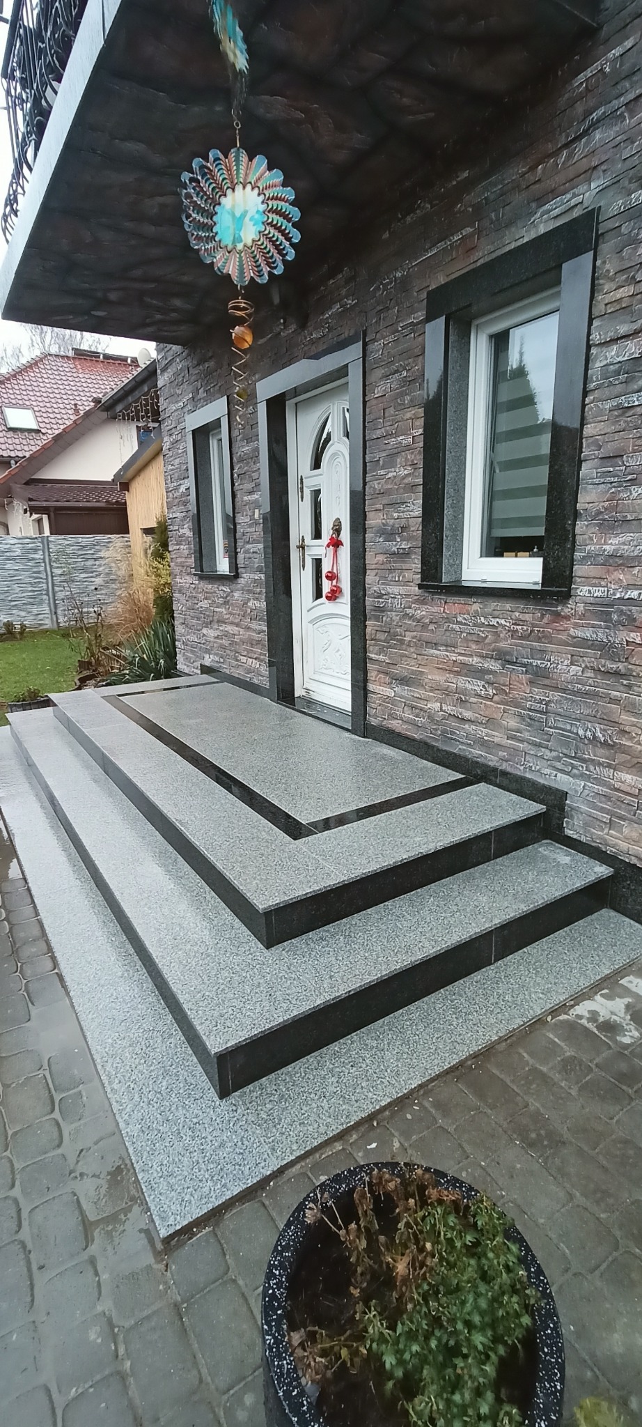 Granitowe schody wejściowe z czarnymi obramowaniami okien i drzwi, dekoracja nad wejściem. Widok na elewację domu z kamienną okładziną i rośliną w donicy.