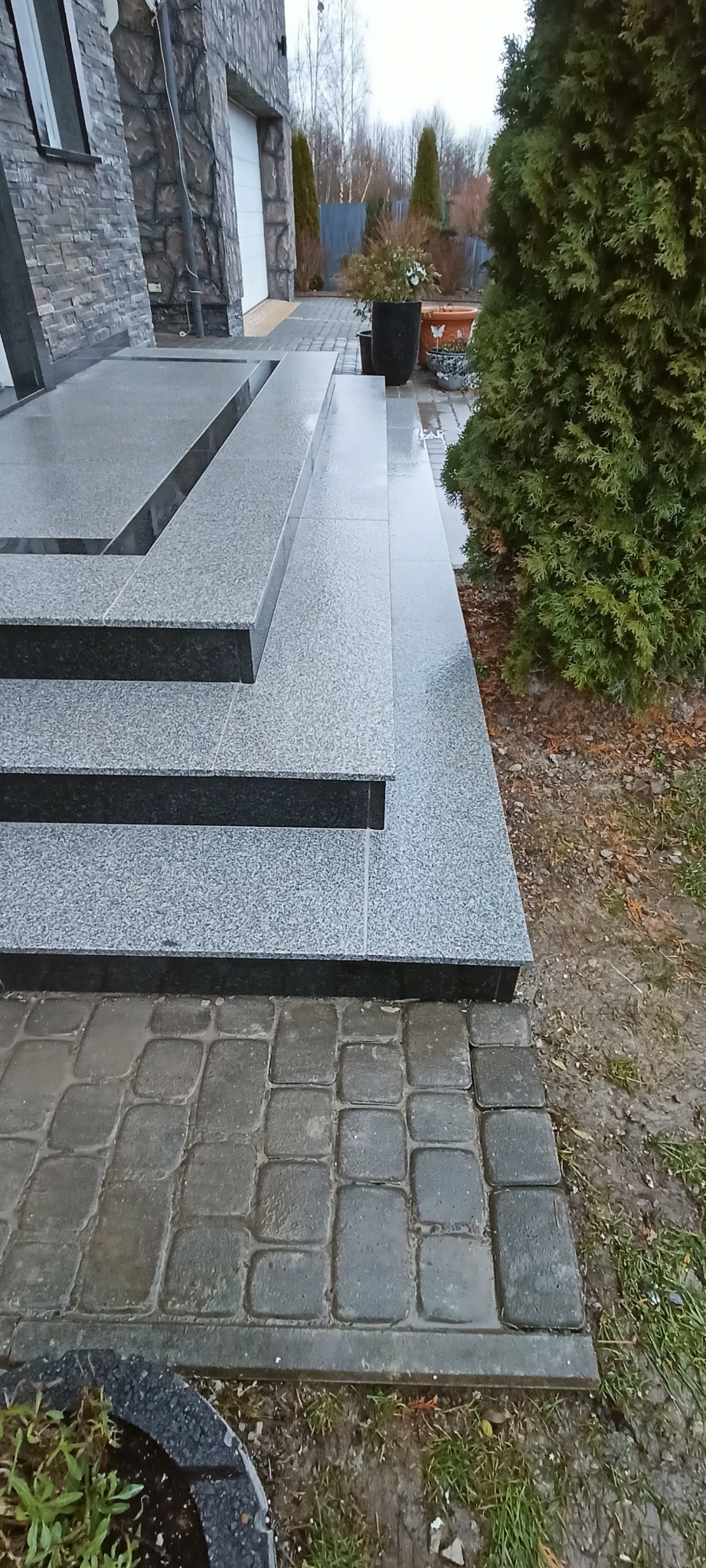 Granitowe schody zewnętrzne z ciemnymi krawędziami prowadzą do domu z kamienną elewacją. Obok rośnie tuja, a w tle widać kostkę brukową i roślinność.