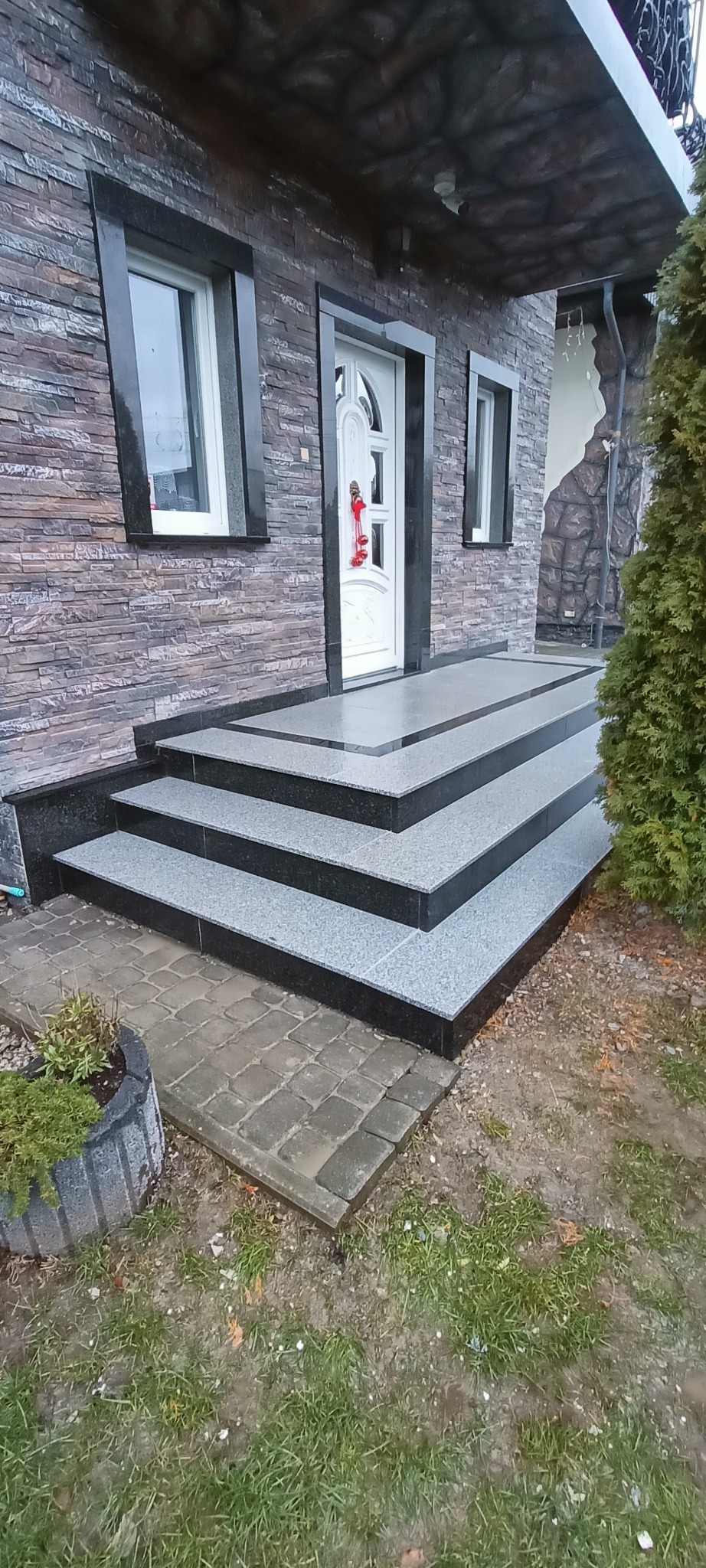 Kamienne schody wejściowe z szarego granitu prowadzą do białych drzwi domu z elewacją z kamienia łupanego. Widoczne czarne obramowania okien i roślinność.