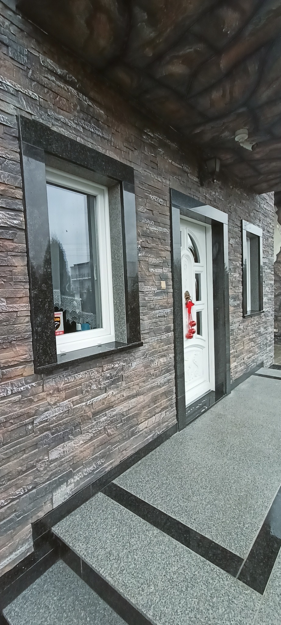 Elewacja budynku z kamienia dekoracyjnego, okna i drzwi obramowane czarnym granitem. Parapet z granitu. Schody wejściowe wyłożone kamieniem. Dekoracja świąteczna na drzwiach.
