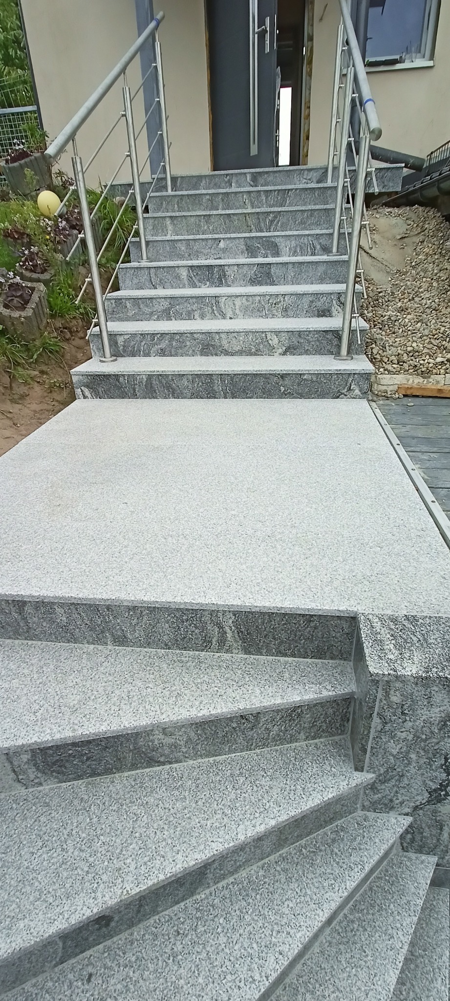 Zewnętrzne schody z jasnoszarego kamienia z metalową balustradą prowadzące do drzwi wejściowych. Widoczne rośliny i kamienie ozdobne w tle.