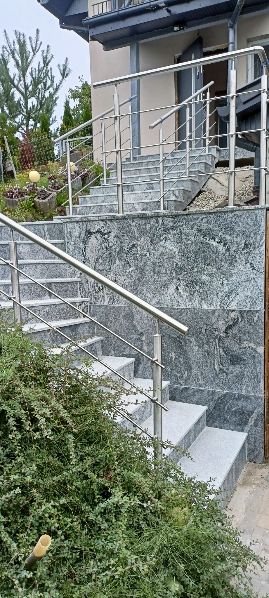 Zewnętrzne schody z szarego kamienia z metalową balustradą prowadzące do wejścia domu. Widoczne elementy ogrodu i elewacja budynku.