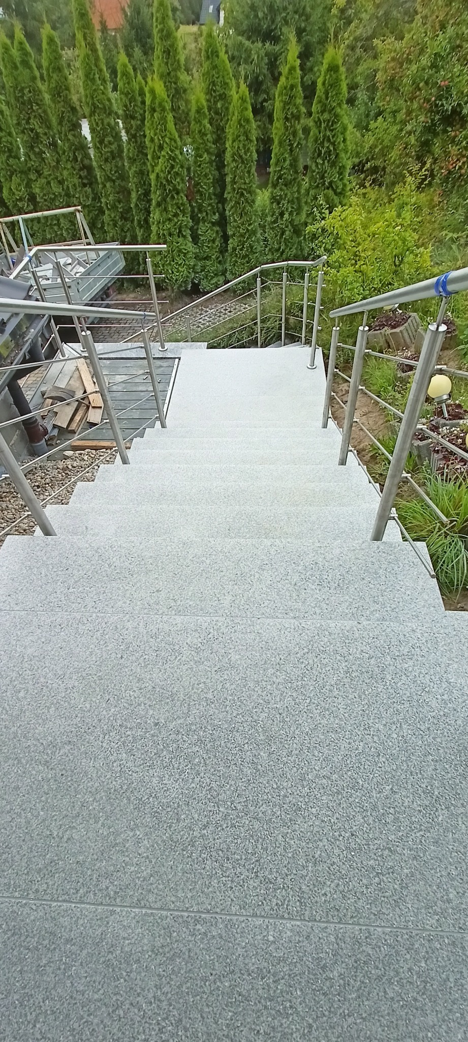 Zewnętrzne schody z jasnoszarego kamienia z metalową balustradą, prowadzące w dół do ogrodu z tujami. Widok z góry, symetryczny kadr.