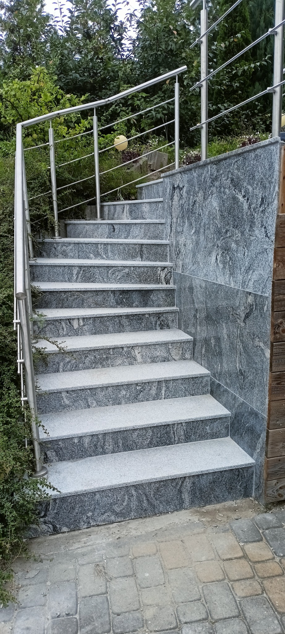 Zewnętrzne schody z szarego kamienia z metalową balustradą, otoczone zielenią. Widoczne detale faktury kamienia i kostki brukowej u podstawy schodów.