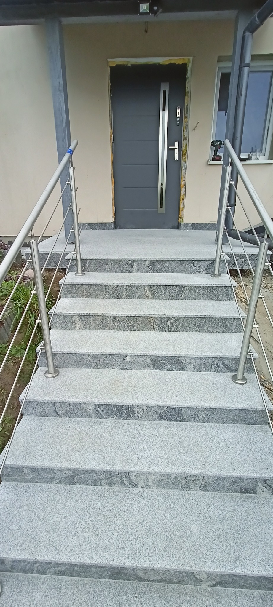 Granitowe schody zewnętrzne z metalową balustradą prowadzące do szarych drzwi wejściowych. Widoczne narzędzia na parapecie okna.