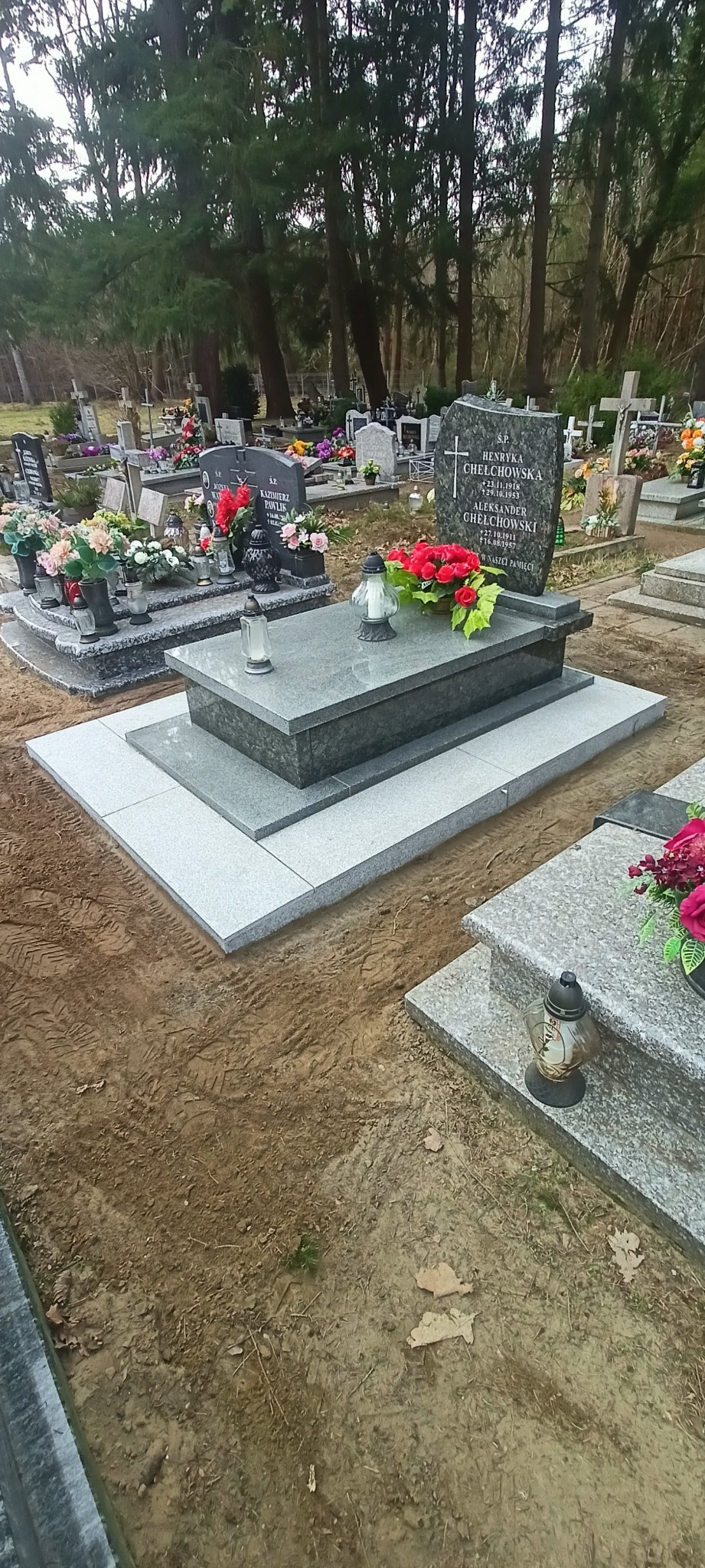 Szary nagrobek z granitu na cmentarzu, z czerwonymi kwiatami i zniczem. Widoczne inne nagrobki i drzewa w tle. Ujęcie z poziomu gruntu.