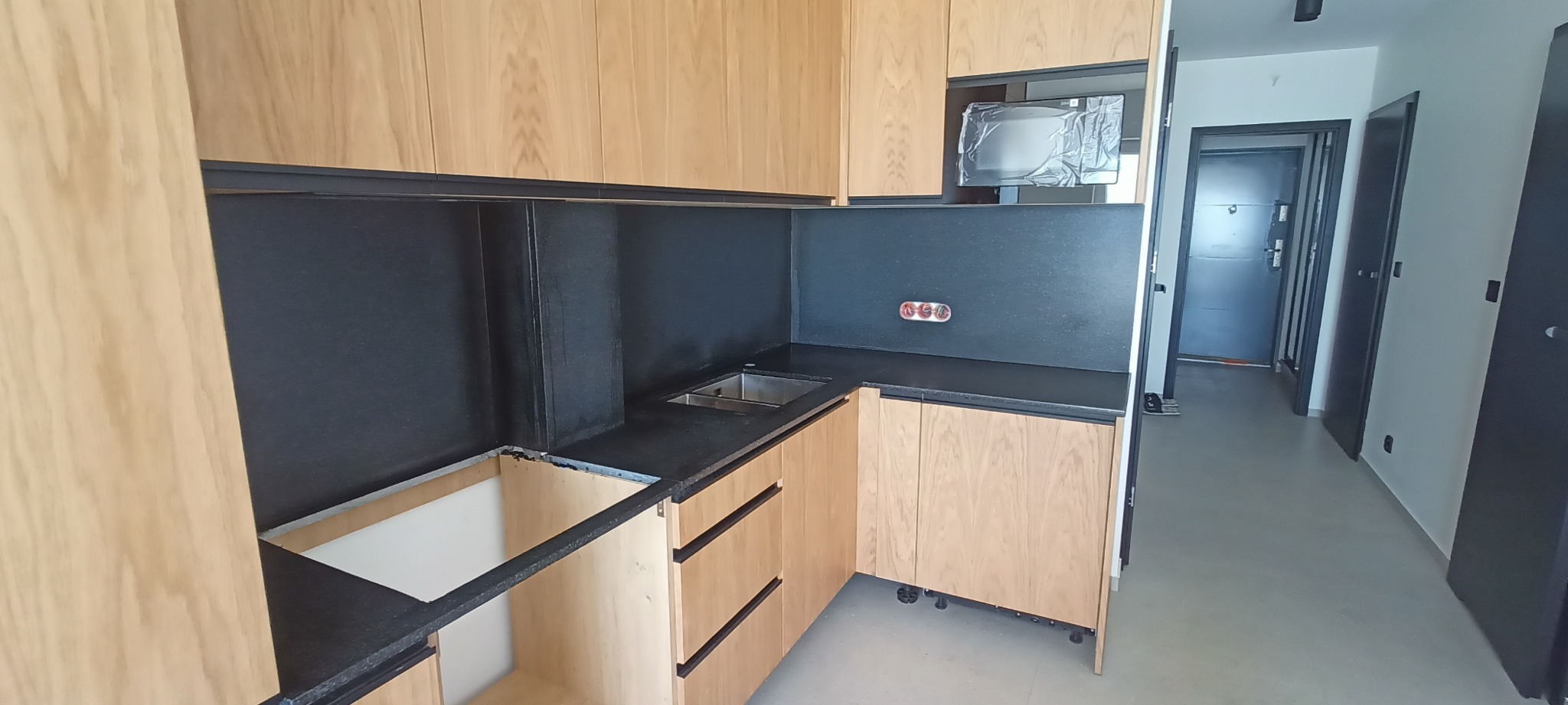 Nowoczesna kuchnia z drewnianymi szafkami i czarnym blatem kamiennym. Widoczny zlew i fragment niezabudowanej szafki. W tle korytarz z drzwiami.