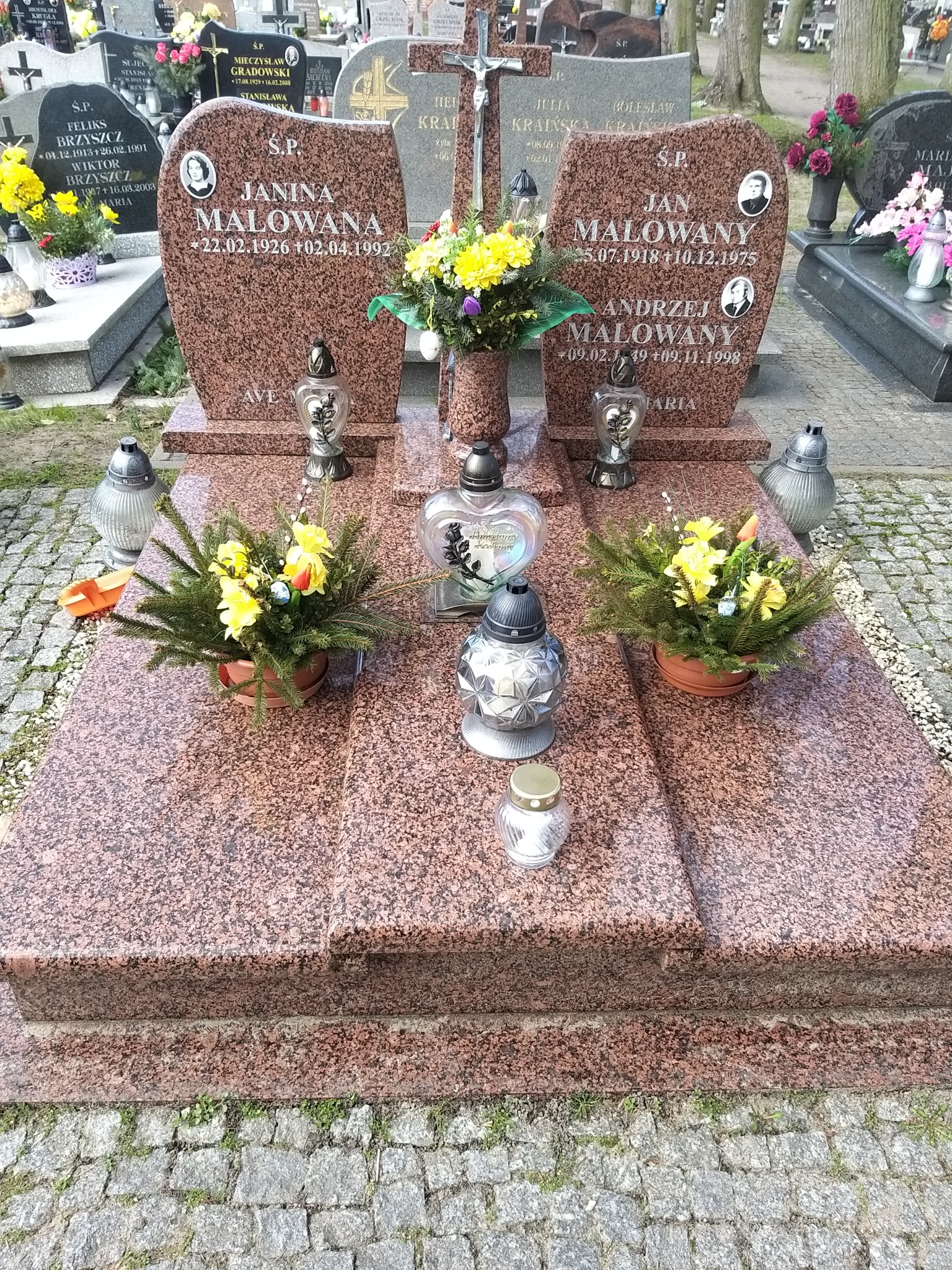 Granitowy pomnik nagrobny z dwoma tablicami, ozdobiony kwiatami w doniczkach i lampionami, na cmentarzu z brukowaną ścieżką.