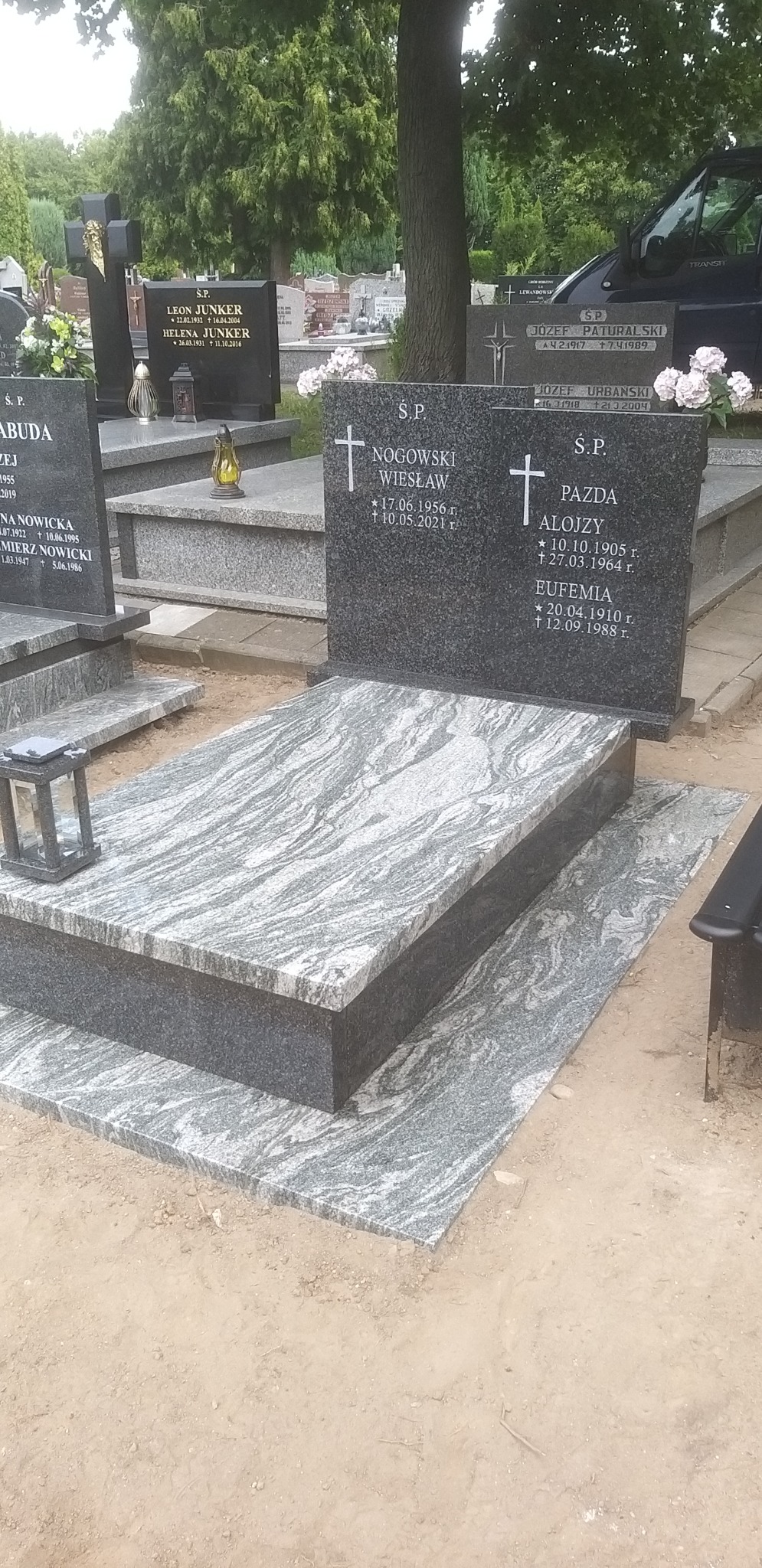 Szary, dwupoziomowy pomnik nagrobny z wzorzystego kamienia, z wyrytymi imionami i datami, na tle cmentarza z innymi nagrobkami i drzewami.