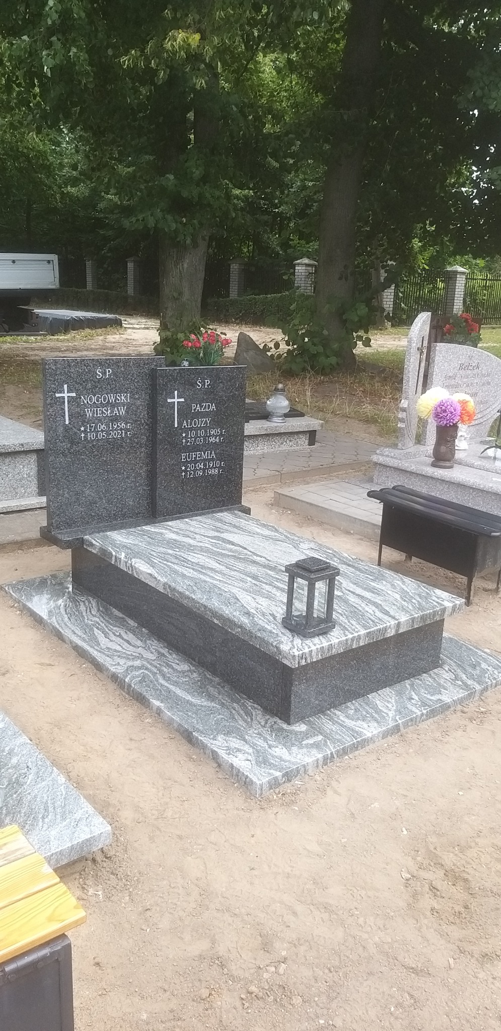 Granitowy nagrobek podwójny z jasnoszarym wzorem żył na płycie, z czarną latarenką, na cmentarzu, otoczony zielenią drzew.