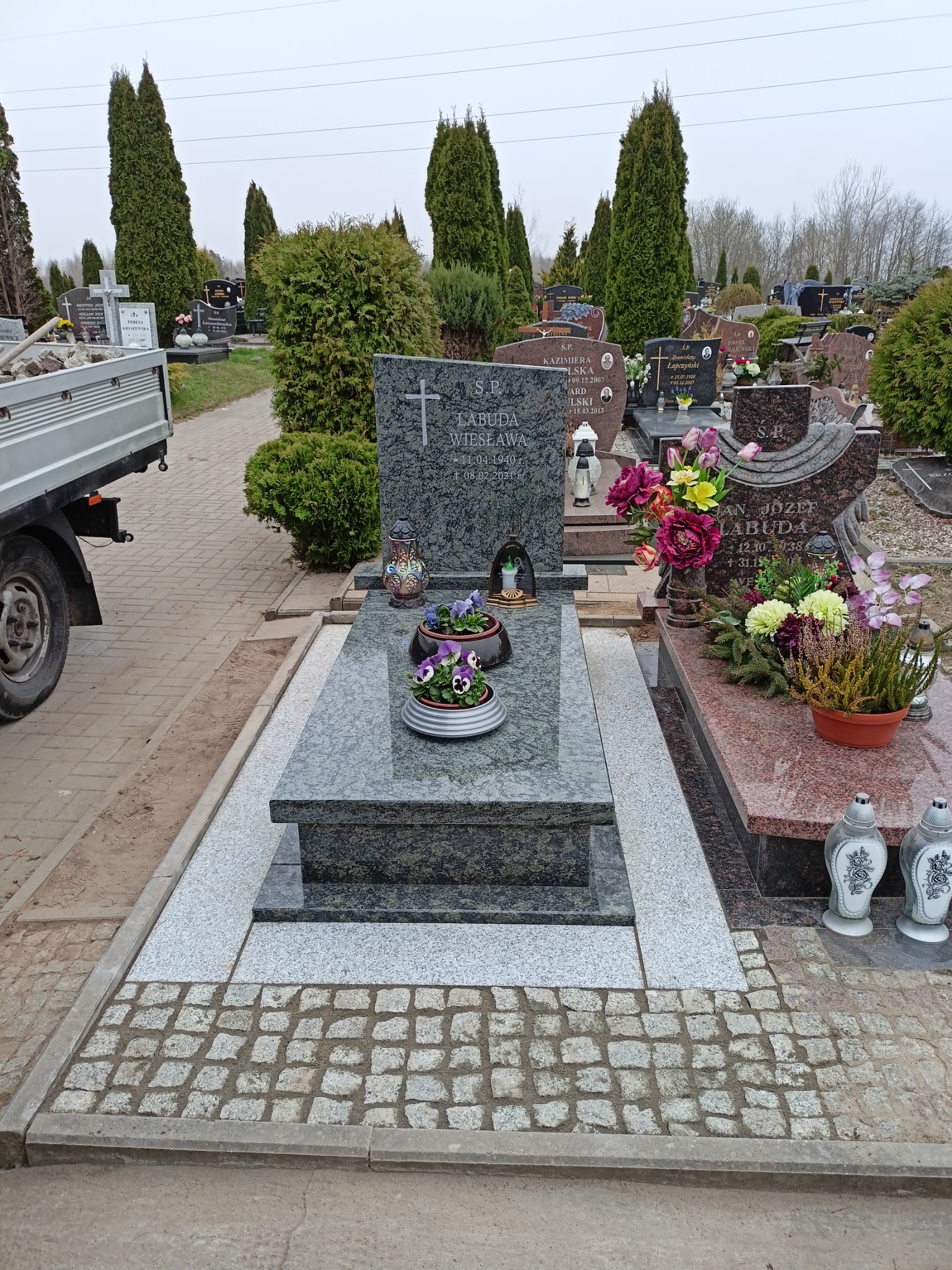 Nowoczesny, szary nagrobek z granitu z ciemniejszymi elementami, z kwiatami w doniczkach, na cmentarzu, częściowo widoczna skrzynia ładunkowa samochodu dostawczego z kamieniami.