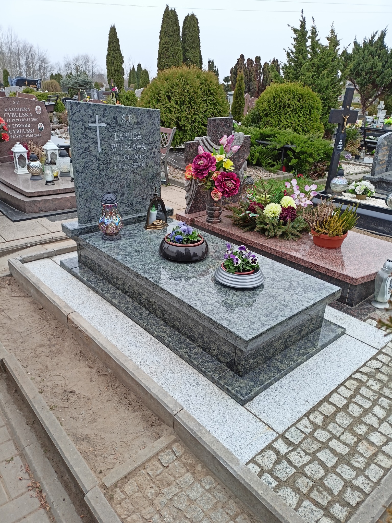 Granitowy pomnik z inskrypcją i krzyżem, ozdobiony kwiatami w doniczkach i lampionem, na cmentarzu z brukowaną ścieżką i zielenią w tle.