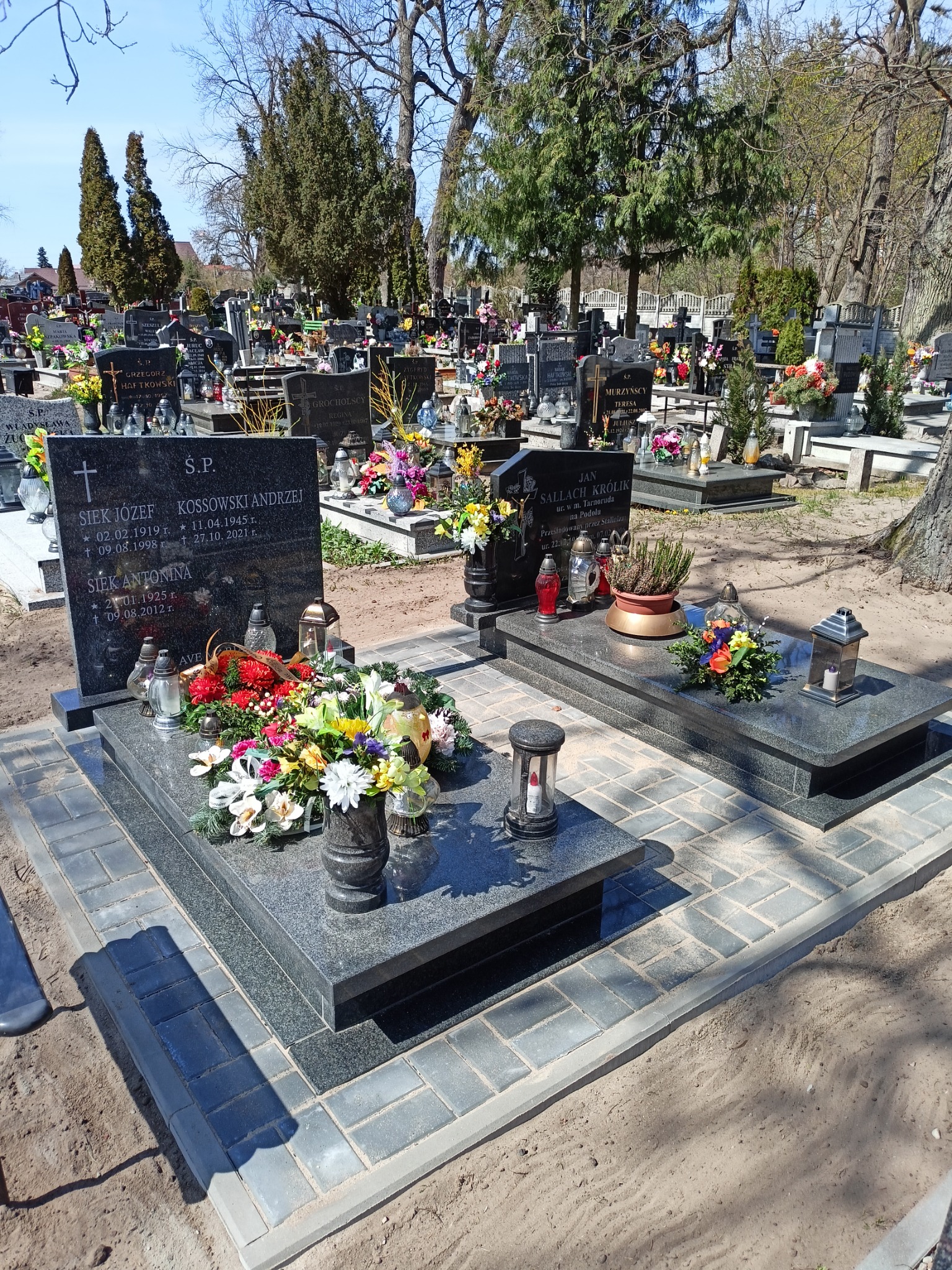 Dwa ciemne, granitowe pomniki na cmentarzu, otoczone szarą kostką brukową, z licznymi kwiatami i lampionami, w tle inne nagrobki i drzewa.