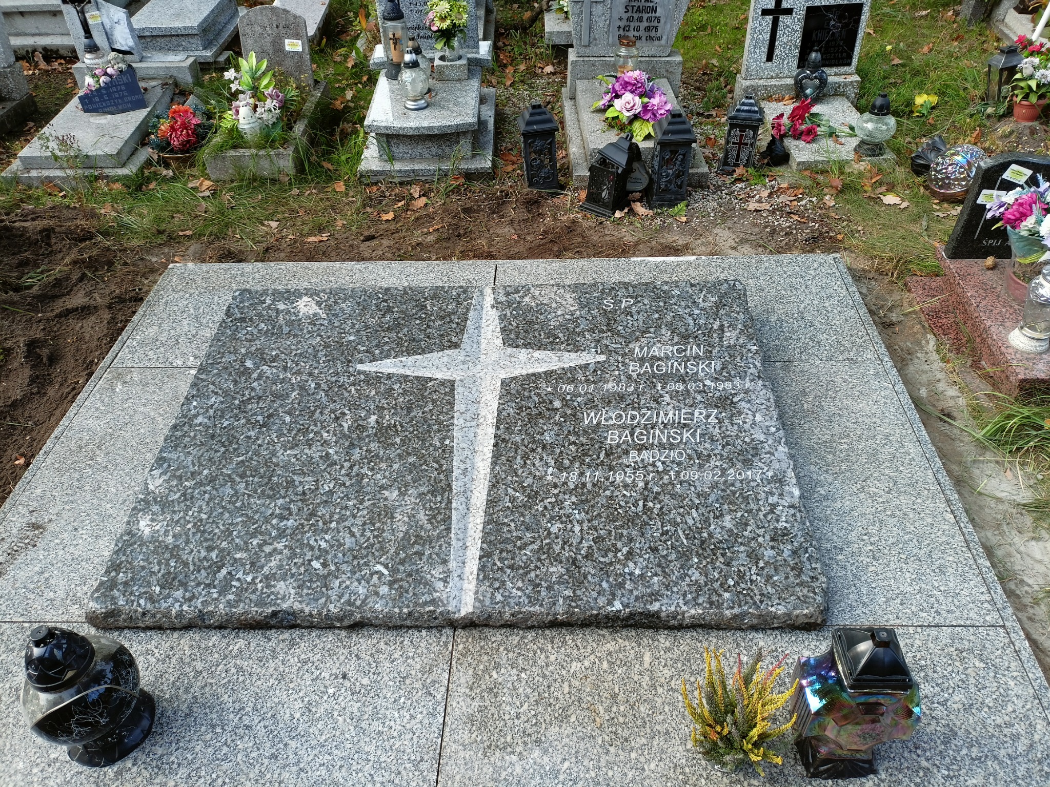 Granitowy nagrobek z wygrawerowanym krzyżem i danymi zmarłych, otoczony innymi nagrobkami i zniczami na cmentarzu.