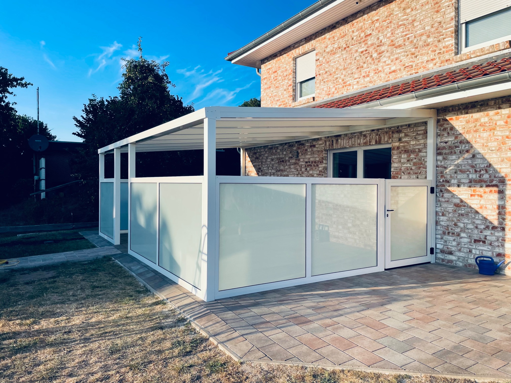 Nowoczesna, biała pergola z mlecznymi szybami, przylegająca do ceglanego budynku, z widocznym fragmentem tarasu z kostki brukowej i błękitnym konewką.