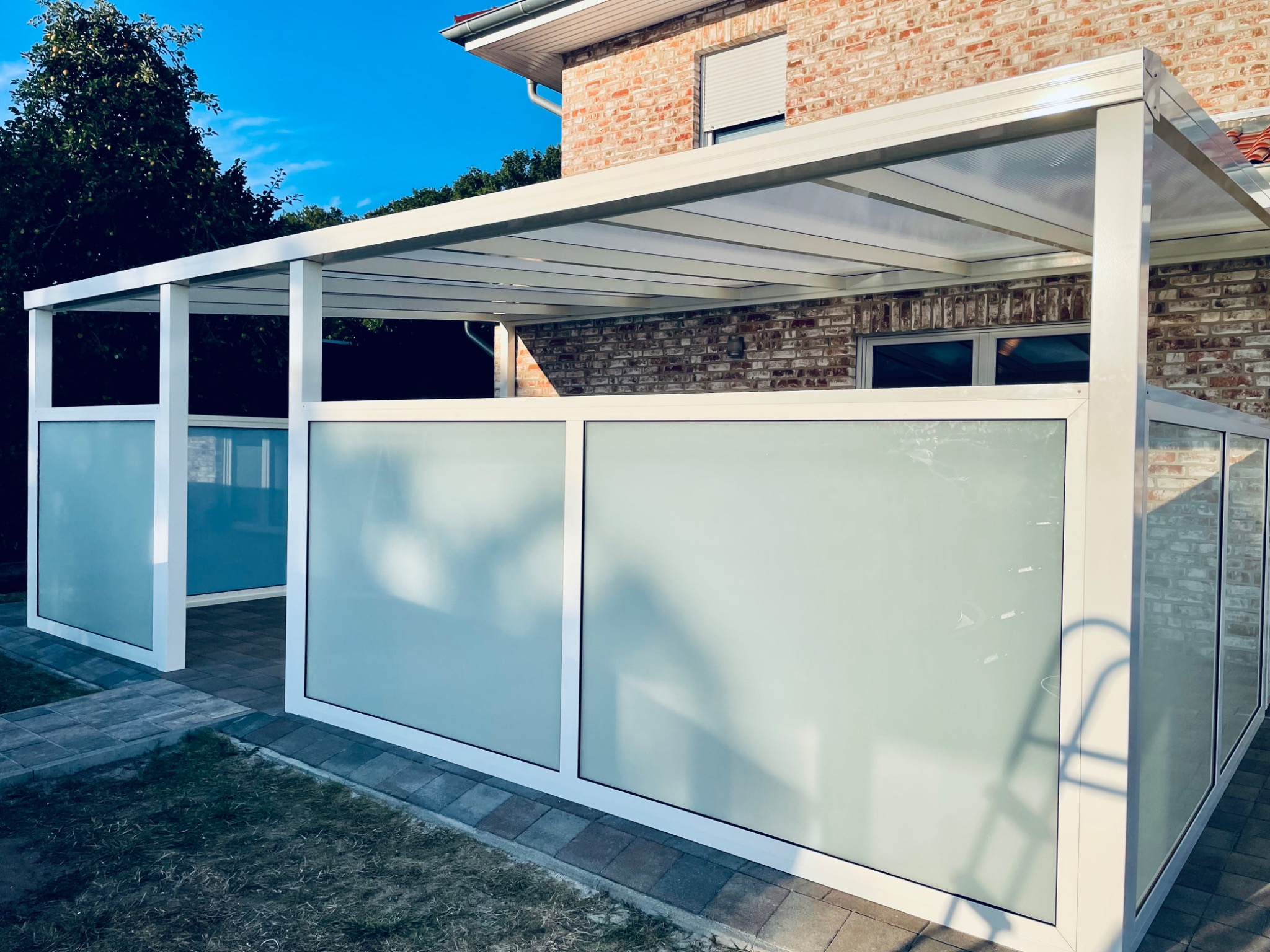 Nowoczesna pergola z białymi ramami i matowymi panelami szklanymi, przylegająca do ceglanego domu, z widocznym fragmentem trawnika i kostki brukowej.
