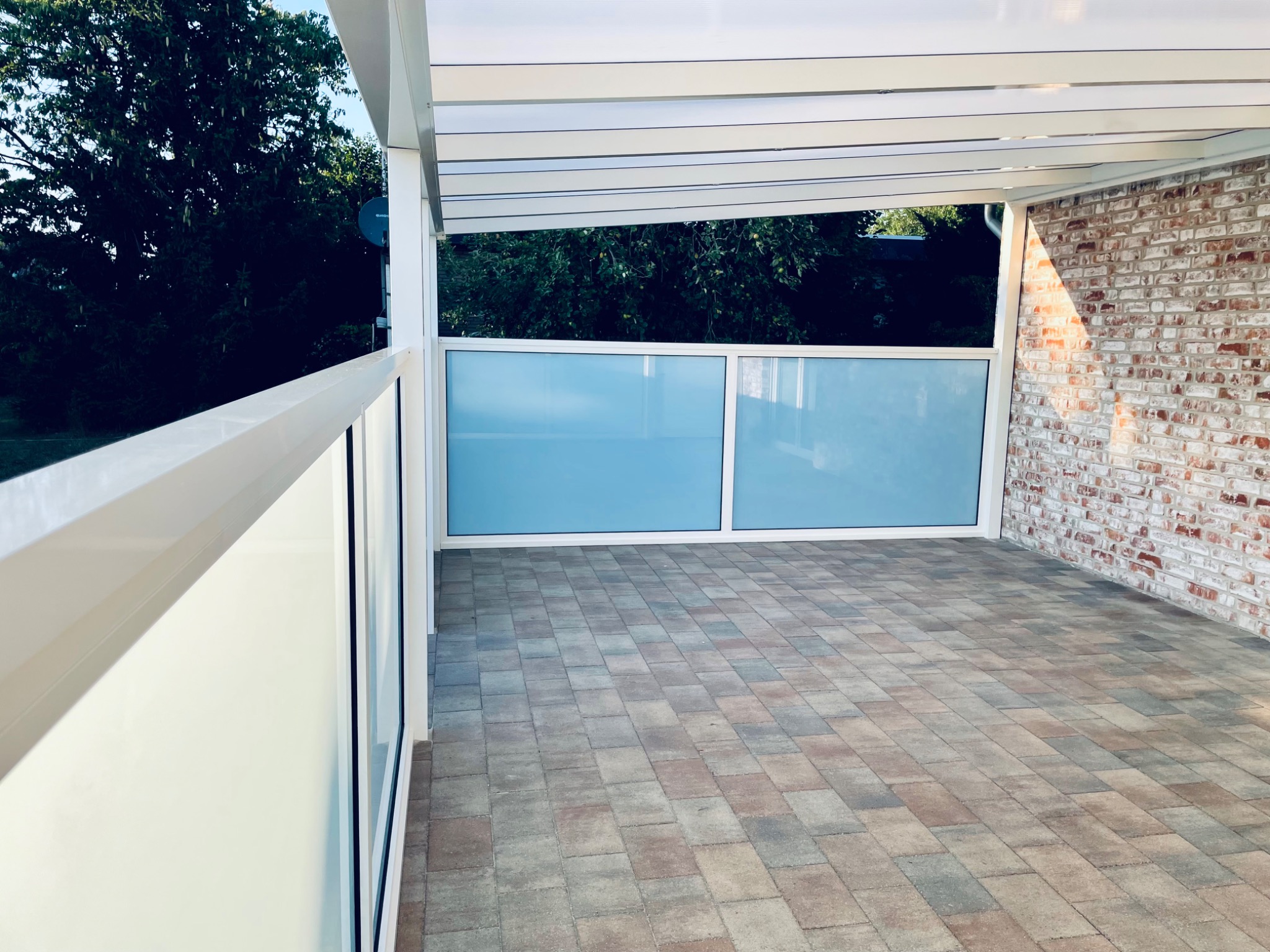 Zadaszony taras z kostki brukowej, częściowo osłonięty szybami, z widoczną ścianą z czerwonej cegły i fragmentem białej pergoli.