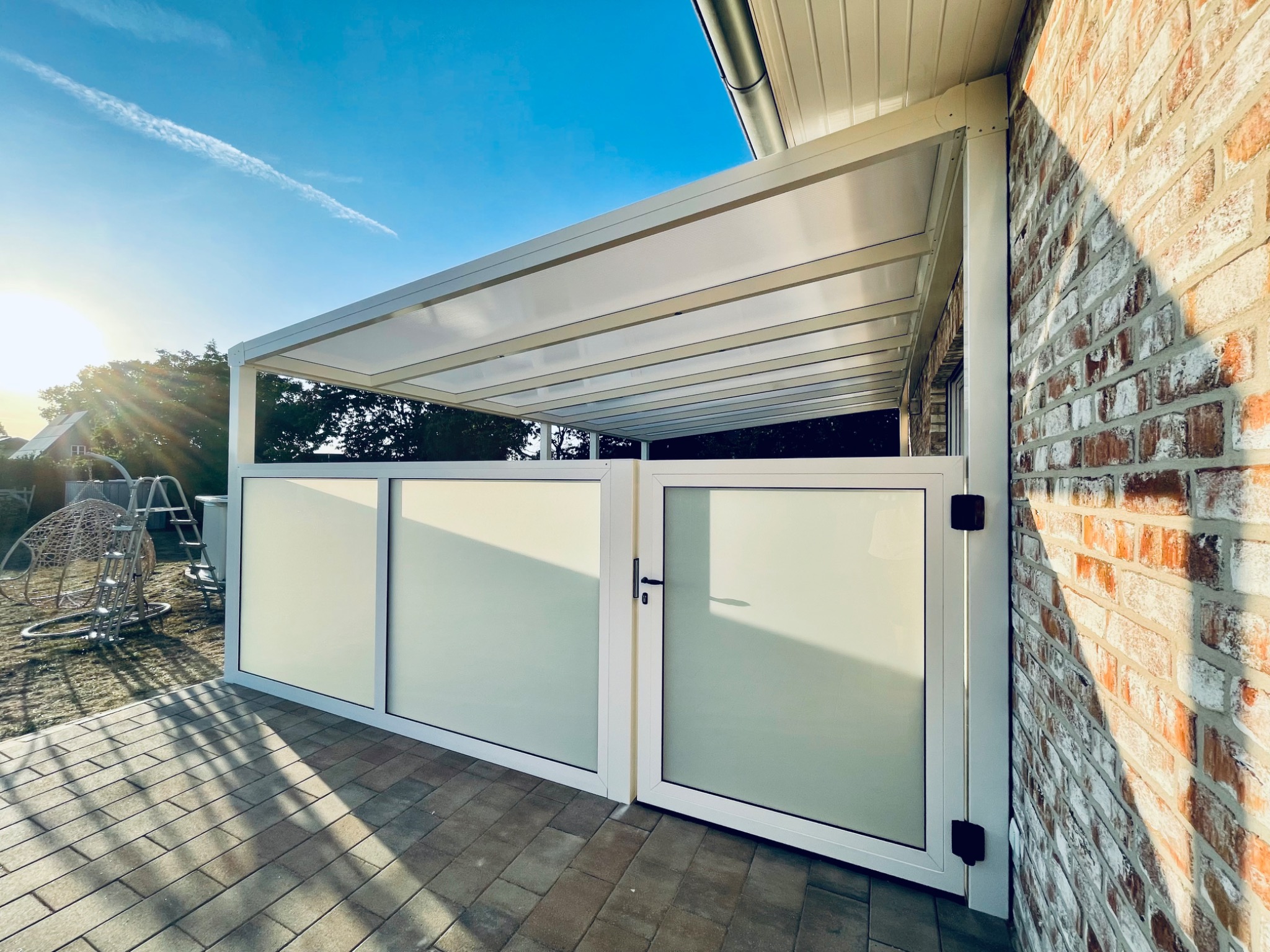 Nowoczesna pergola z białymi, matowymi panelami bocznymi, przylegająca do ceglanego budynku, na wyłożonym kostką brukową tarasie w słoneczny dzień.