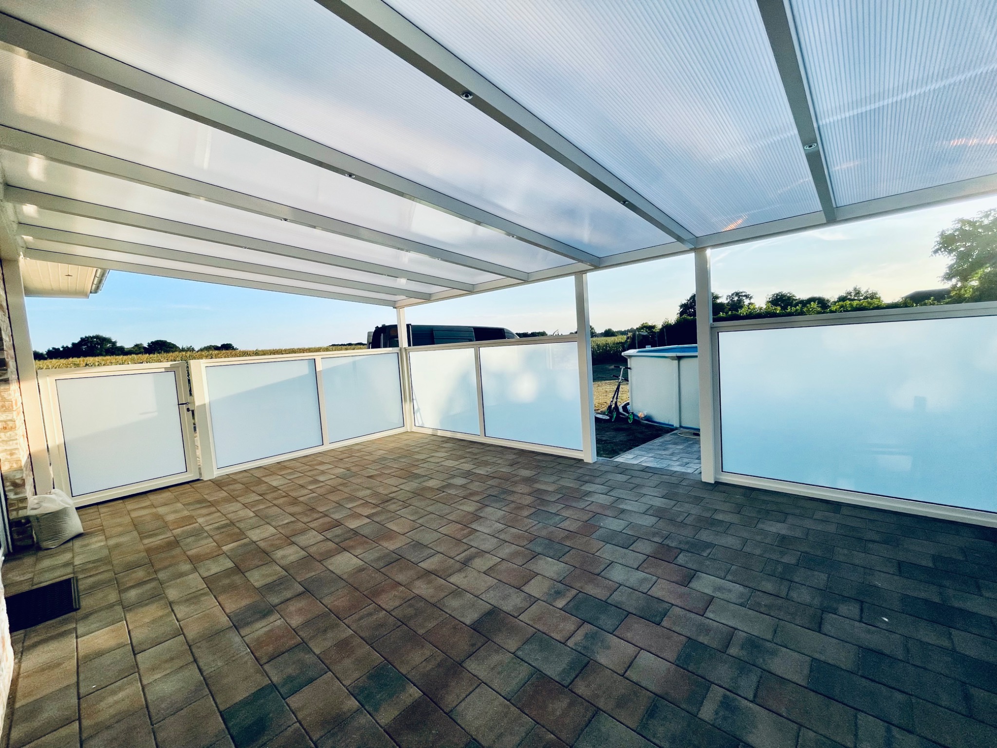 Zadaszony taras z poliwęglanowym dachem i matowymi panelami bocznymi, wyłożony kostką brukową w odcieniach brązu i szarości, z widokiem na ogród i basen w tle.