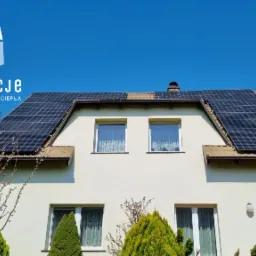 Dom jednorodzinny z zamontowanymi panelami fotowoltaicznymi na dachu, widoczne krzewy ozdobne w ogrodzie, błękitne niebo w tle, logo firmy instalacyjnej w lewym górnym rogu.