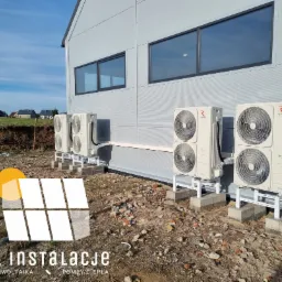 Zewnętrzne jednostki czterech pomp ciepła marki Rotenso Unico, zamontowane na betonowych fundamentach i metalowych stelażach przy hali produkcyjnej z szarej blachy trapezowej, widoczne przewody...