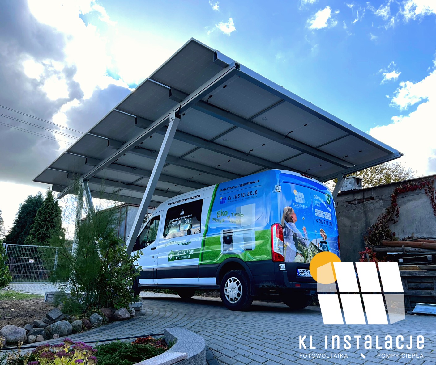 Instalacja fotowoltaiczna Carport z panelami zamontowanymi nad miejscem parkingowym, obok zaparkowany firmowy bus z reklamą usług instalacyjnych, widok na podjazd z kostki brukowej i fragment ogrodu.