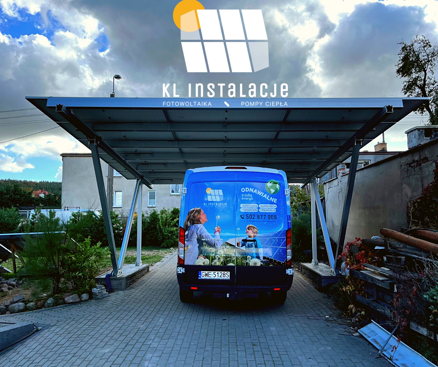 Carport z panelami fotowoltaicznymi, widok spod konstrukcji na zaparkowany samochód dostawczy z reklamą firmy instalującej odnawialne źródła energii, błękitne niebo z chmurami w tle.