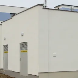 Zewnętrzna stacja transformatorowa z dwoma parami metalowych drzwi oznaczonych tabliczkami informacyjnymi, przylegająca do budynku mieszkalnego z widocznym fragmentem elewacji w kolorze białym...