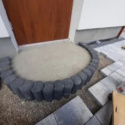 Przygotowanie podłoża pod wejście do domu: ułożona obwódka z ciemnych, okrągłych elementów betonowych otacza wysypaną warstwę piasku, obok ułożone szare płyty chodnikowe, widoczny worek z zaprawą...