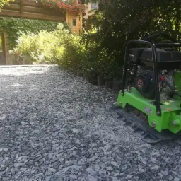 Zielona zagęszczarka spalinowa ubija szarą podsypkę kamienną na podwórku z elementami ogrodu w tle.