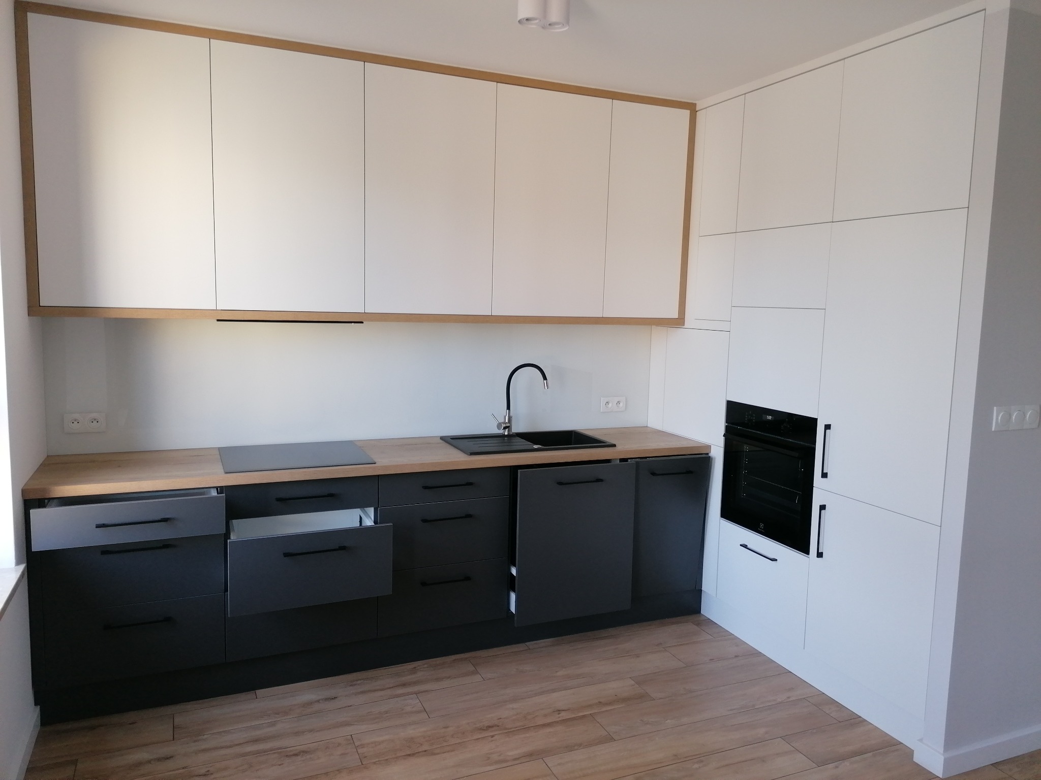 Nowoczesna kuchnia z białymi górnymi szafkami obramowanymi drewnianym dekorem i ciemnoszarymi dolnymi szafkami z otwartymi szufladami, blat z płyty laminowanej w kolorze drewna, czarny zlew i płyta...