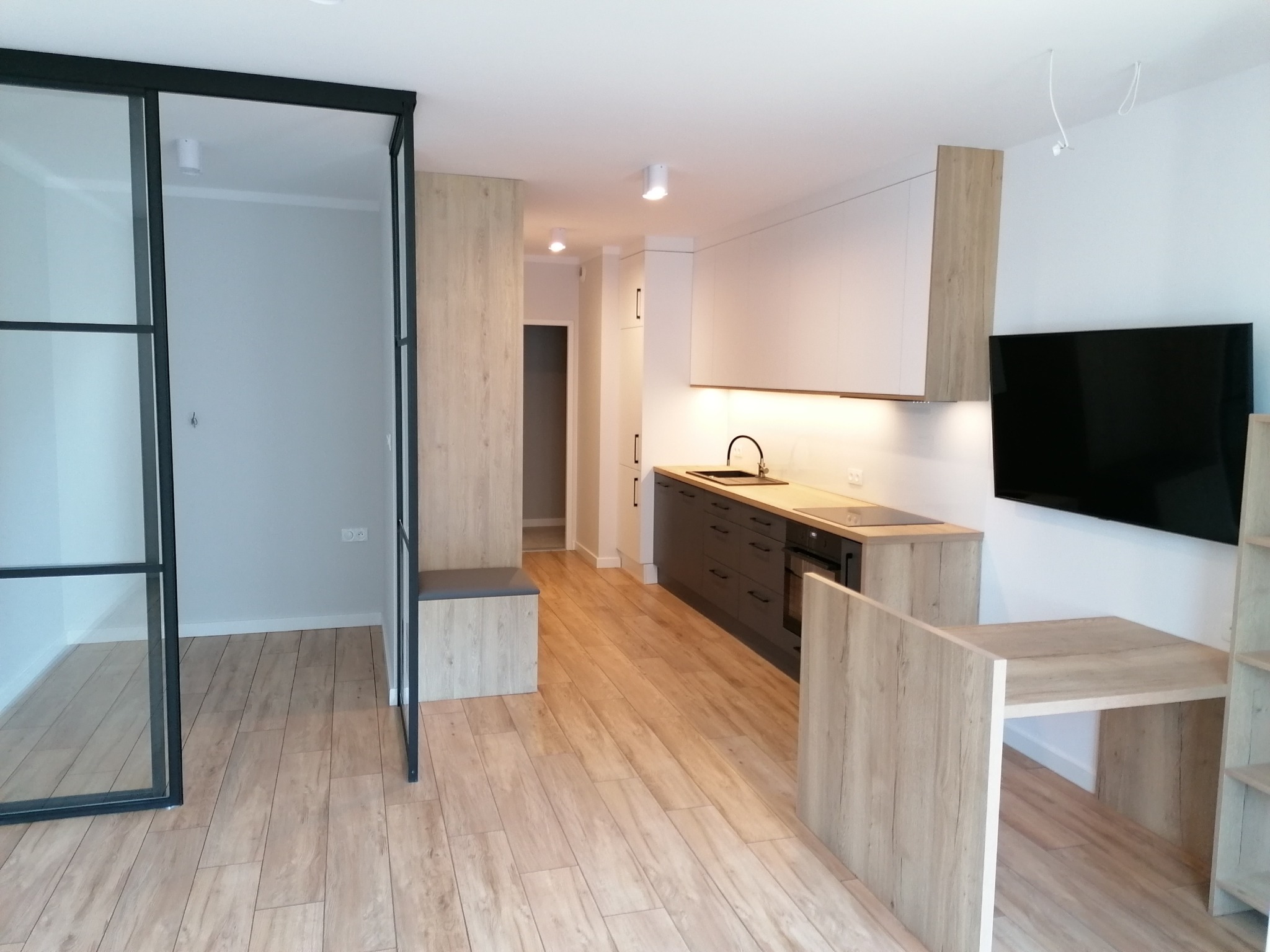 Nowoczesna kuchnia z frontami i blatem z płyty laminowanej w odcieniach szarości i drewna, z widocznym fragmentem szklanej ścianki działowej z czarną ramą oraz drewnianą podłogą.