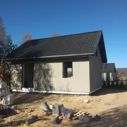 Surowy dom jednorodzinny z ciemną dachówką w trakcie budowy, widoczne bloczki betonowe i otwory okienne bez okien, na tle błękitnego nieba i krajobrazu.
