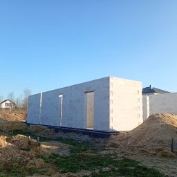 Wznoszone ściany domu z betonu komórkowego na fundamencie, widoczne otwory okienne i drzwiowe, hałdy piasku, w tle dom jednorodzinny i błękitne niebo.