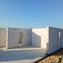 Wnętrze domu w trakcie budowy, ściany z białych bloczków, widoczne otwory okienne i drzwiowe, betonowa podłoga, błękitne niebo w tle. Surowy stan budynku.