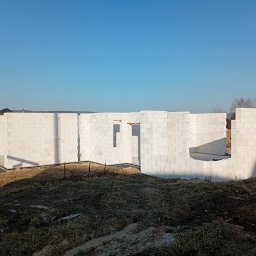 Wznoszenie ścian domu z białych bloczków betonu komórkowego. Widoczne otwory okienne i drzwiowe. Fundamenty i początek ścian na tle błękitnego nieba.