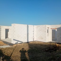 Wznoszenie ścian parteru z białych bloczków betonu komórkowego na fundamencie. Widoczny cień osoby fotografującej. Błękitne niebo w tle.