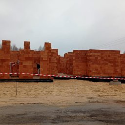 Salwa Budownictwo - Wznoszenie ścian z czerwonej cegły na budowie domu. Widoczny pracownik wyrównujący piasek wewnątrz fundamentów, otoczonych taśmą ostrzegawczą.