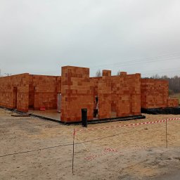 Salwa Budownictwo - Wznoszenie ścian domu z czerwonej cegły ceramicznej. Widoczne fundamenty i izolacja. Na pierwszym planie piasek i taśma ostrzegawcza. Pochmurne niebo w tle.