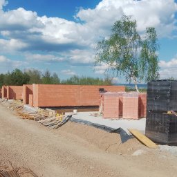 Salwa Budownictwo - Budowa szeregówki z czerwonej cegły. Częściowo wzniesione ściany, palety z cegłami, teren budowy w słoneczny dzień. Widoczni pracownicy budowlani.