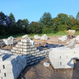 Salwa Budownictwo - Wznoszenie ścian fundamentowych z bloczków betonowych na budowie domu jednorodzinnego. Widoczne zbrojenie i palety z bloczkami. W tle zieleń drzew i błękitne niebo.
