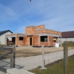 Salwa Budownictwo - Budowa domu jednorodzinnego w stanie surowym otwartym, z widocznymi bloczkami ceramicznymi i betonowymi nadprożami, na tle błękitnego nieba i sąsiednich budynków.